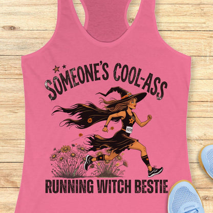 A pink racerback tank top laid flat on a light wood surface, featuring a graphic of a woman running as a witch with the text 'SOMEONE'S COOL-ASS RUNNING WITCH BESTIE'. Partial blue shoes are visible at the edges.