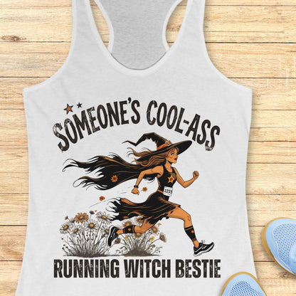 A white racerback tank top laid flat on a light wood background, featuring a graphic of a woman running as a witch with the text 'SOMEONE'S COOL-ASS RUNNING WITCH BESTIE'. Partial blue shoes are visible at the bottom right.