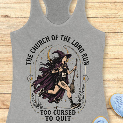 A heather gray racerback tank top featuring a graphic of a witch running with a broomstick, surrounded by the text 'THE CHURCH OF THE LONG RUN TOO CURSED TO QUIT'. The tank top is displayed flat on a light wood background with a pair of blue sneakers partially visible in the corner.