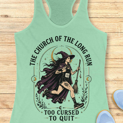 A mint green racerback tank top laid flat on a light wood background, featuring a large graphic of a witch running with a broomstick and the text 'THE CHURCH OF THE LONG RUN TOO CURSED TO QUIT'.