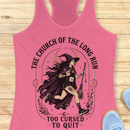 A pink racerback tank top laid flat on a light wood surface, featuring a large graphic of a witch running with a broomstick and the text 'THE CHURCH OF THE LONG RUN TOO CURSED TO QUIT.' Partial blue shoes are visible at the edges as styling props.