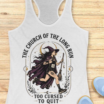 A white racerback tank top with a graphic of a witch riding a broomstick, surrounded by the text 'THE CHURCH OF THE LONG RUN TOO CURSED TO QUIT'. The tank top is displayed flat on a light wood background with blue shoes partially visible in the corner.