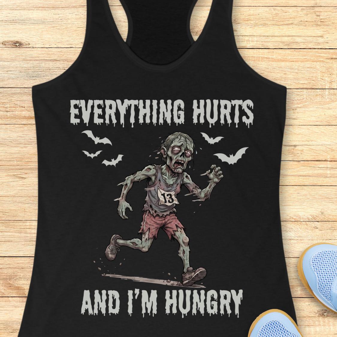 A black racerback tank top laid flat on a light wood surface, featuring a zombie running graphic with the text 'EVERYTHING HURTS AND I'M HUNGRY' in white distressed lettering. A partial blue shoe is visible at the bottom right as a styling prop.