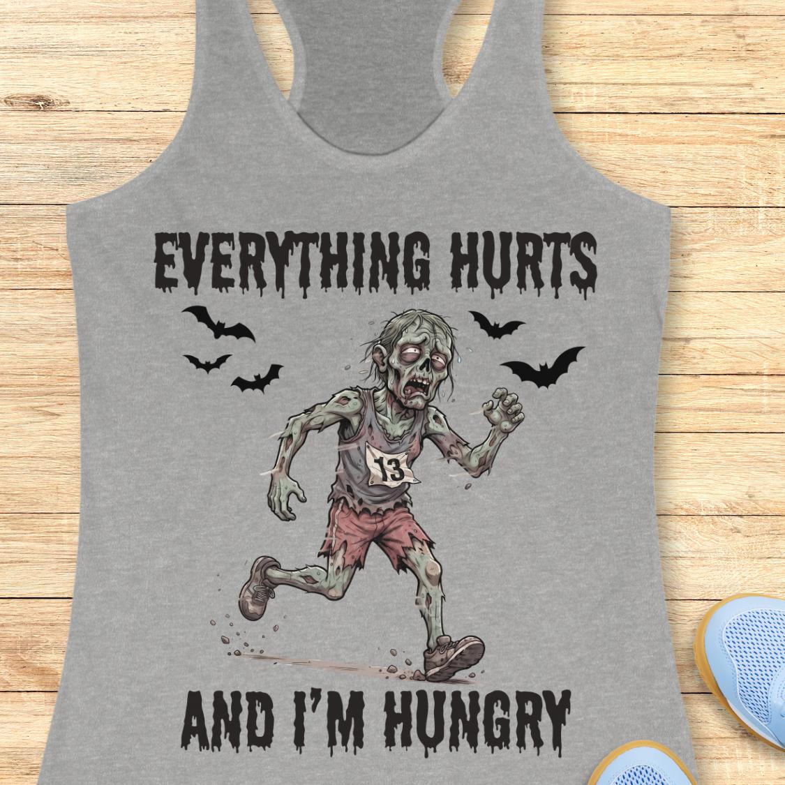 A heather gray racerback tank top laid flat on a light wood background, featuring a zombie graphic with the text 'EVERYTHING HURTS AND I'M HUNGRY' and bat silhouettes.