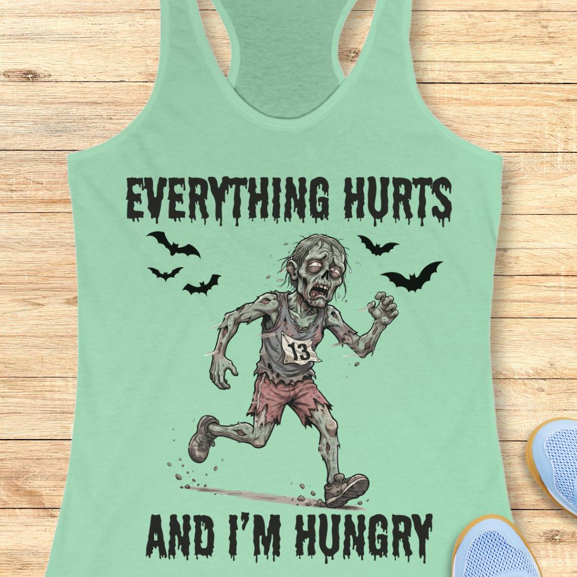 A mint green racerback tank top laid flat on a light wood background, featuring a zombie running graphic with the text 'EVERYTHING HURTS AND I'M HUNGRY' and small bat illustrations.