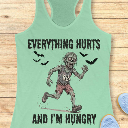 A mint green racerback tank top laid flat on a light wood background, featuring a zombie running graphic with the text 'EVERYTHING HURTS AND I'M HUNGRY' and small bat illustrations.