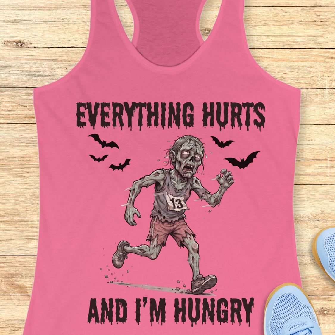 A pink racerback tank top laid flat on a light wood surface, featuring a zombie graphic with the text 'EVERYTHING HURTS AND I'M HUNGRY' and small bat silhouettes.