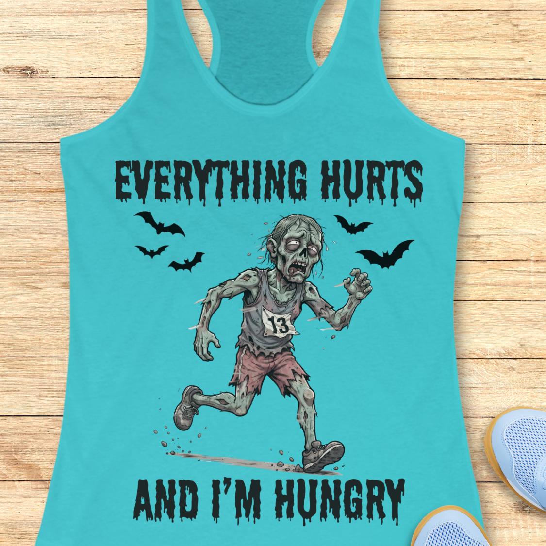A turquoise racerback tank top laid flat on a light wood surface, featuring a zombie graphic with the text 'EVERYTHING HURTS AND I'M HUNGRY' and small bat silhouettes. Part of a blue shoe is visible at the lower right as a styling prop.