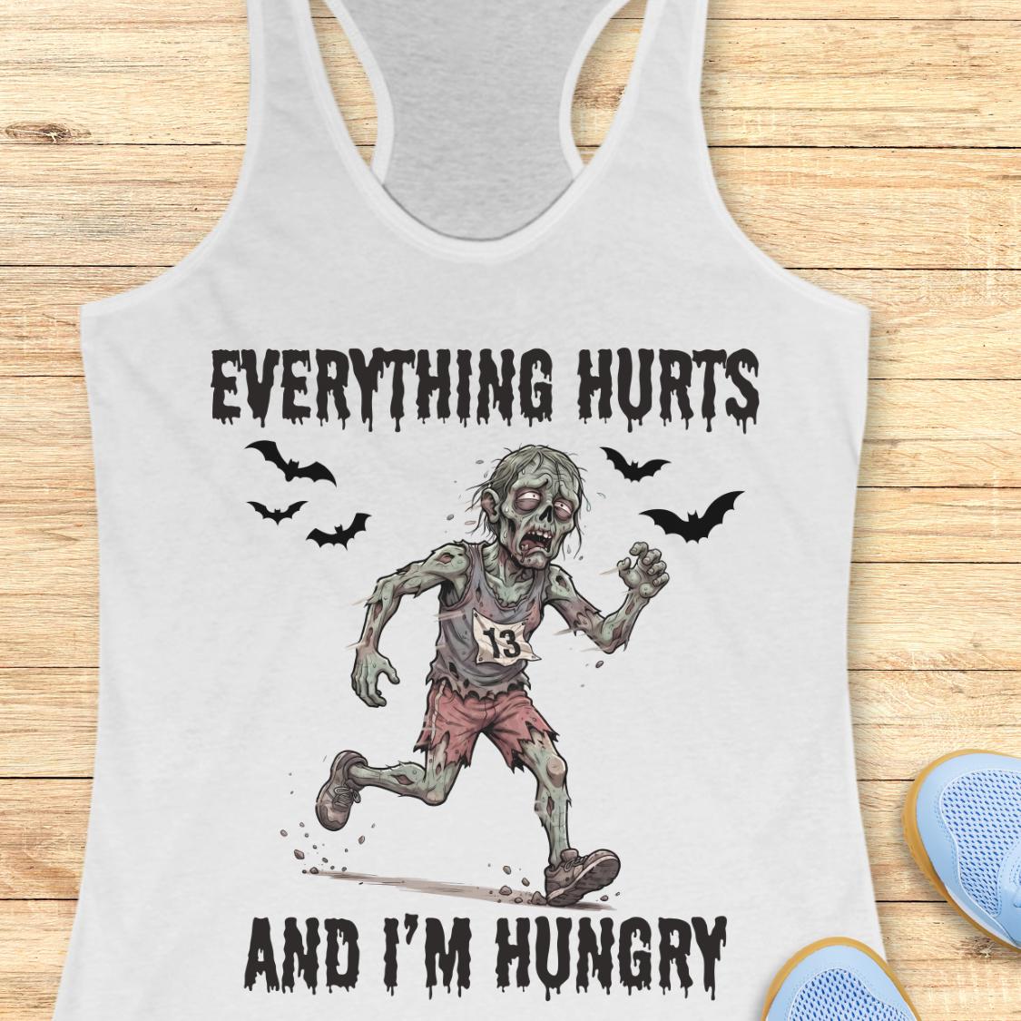 A white racerback tank top laid flat on a light wood background, featuring a zombie running graphic with the text 'EVERYTHING HURTS AND I'M HUNGRY' and small bat silhouettes.