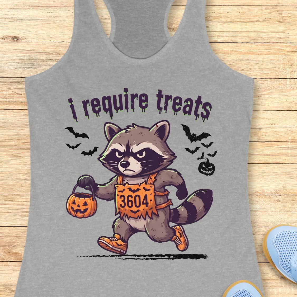 A heather gray racerback tank top featuring a cartoon raccoon in a Halloween costume holding a pumpkin bucket, with the text 'i require treats' above it, displayed on a light wood background.