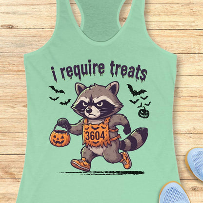 A mint green racerback tank top laid flat on a light wood background, featuring a cartoon raccoon dressed in an orange costume holding a jack-o'-lantern bucket with the text 'i require treats'.
