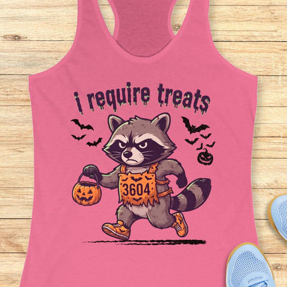 A pink racerback tank top laid flat on a light wood surface, featuring a cartoon raccoon in a Halloween costume holding a pumpkin bucket with the text 'i require treats' above it. Partial blue shoes are visible at the bottom right corners.