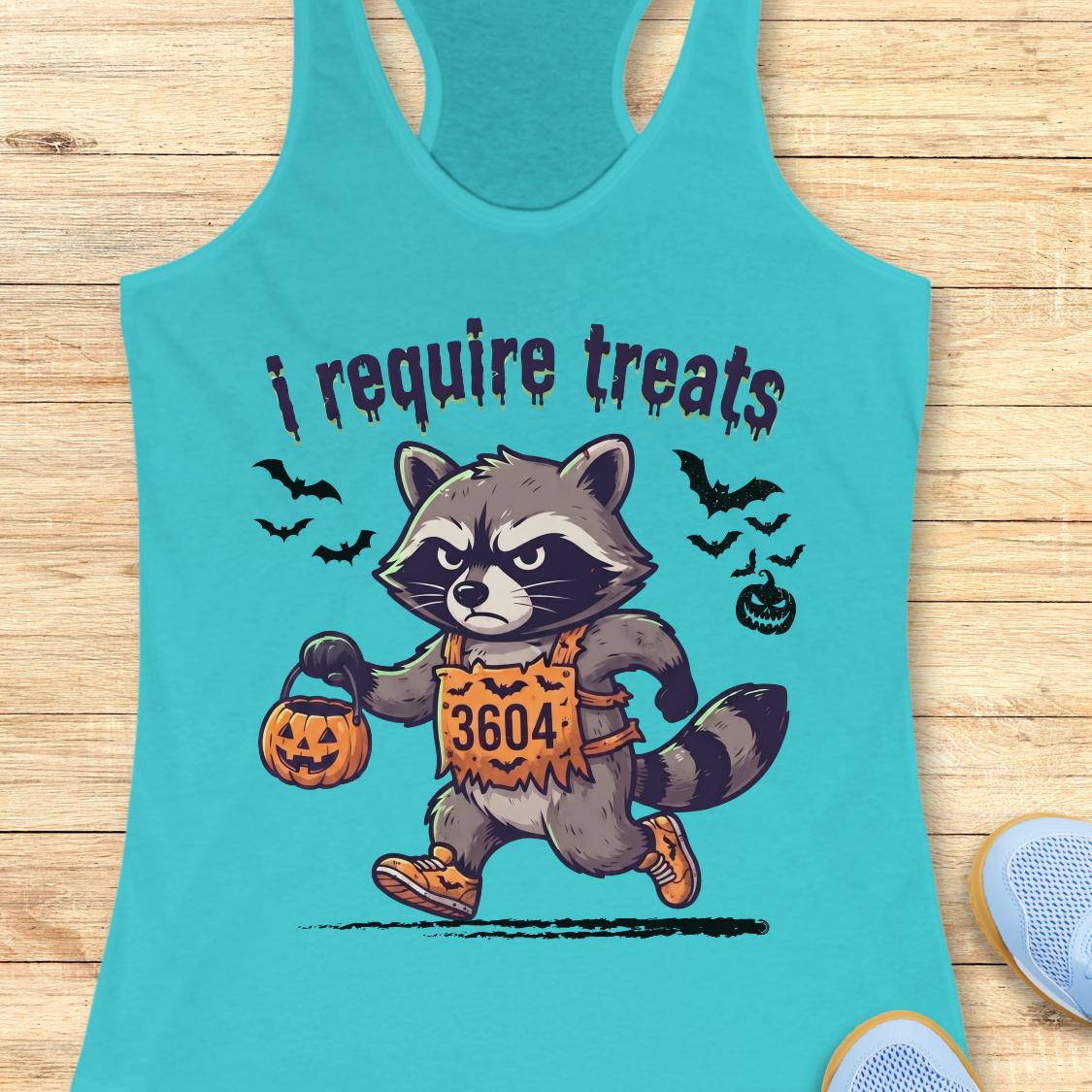 A turquoise racerback tank top laid flat on a light wood surface, featuring a cartoon raccoon in a Halloween costume holding a pumpkin bucket with the text 'i require treats'.