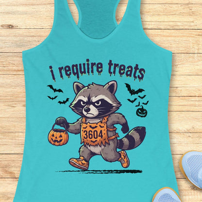 A turquoise racerback tank top laid flat on a light wood surface, featuring a cartoon raccoon in a Halloween costume holding a pumpkin bucket with the text 'i require treats'.