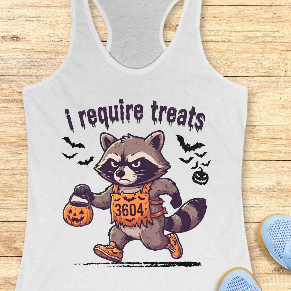 A white racerback tank top laid flat on a light wood background, featuring a cartoon raccoon dressed in an orange Halloween costume with the text 'i require treats' above it. Partial blue shoes are visible at the bottom right as styling props.