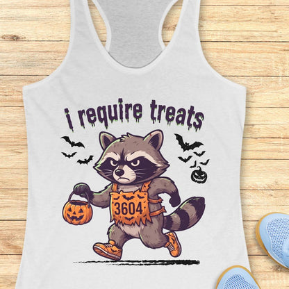 A white racerback tank top laid flat on a light wood background, featuring a cartoon raccoon dressed in an orange Halloween costume with the text 'i require treats' above it. Partial blue shoes are visible at the bottom right as styling props.
