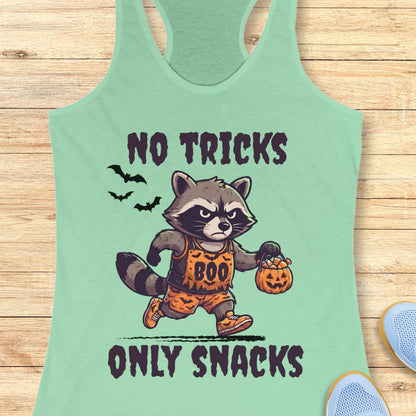A mint green racerback tank top laid flat on a light wood background, featuring a cartoon raccoon in an orange 'BOO' costume holding a jack-o'-lantern, with the text 'NO TRICKS ONLY SNACKS'.