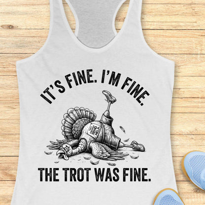 A white racerback tank top laid flat on a light wood background, featuring a black graphic of a lying turkey with one leg raised and the text 'IT'S FINE. I'M FINE.