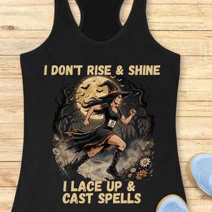 A black racerback tank top laid flat on a light wood surface, featuring a Halloween-themed graphic of a witch running in front of a full moon with the text 'I DON'T RISE & SHINE I LACE UP & CAST SPELLS'. A blue shoe is partially visible at the bottom right.