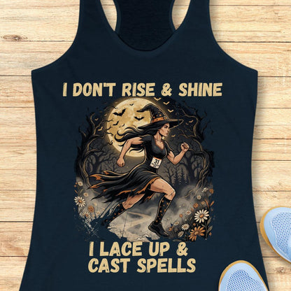 A navy blue racerback tank top laid flat on a light wood background, featuring a Halloween-themed graphic of a witch running under a full moon with the text 'I DON'T RISE & SHINE I LACE UP & CAST SPELLS'.