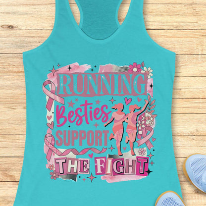 A blue racerback tank top with a pink and black graphic that reads 'running besties support the fight' in stylized lettering, surrounded by pink ribbon motifs and small runner silhouettes, shown on a wooden plank background with partial blue shoes at the edges.