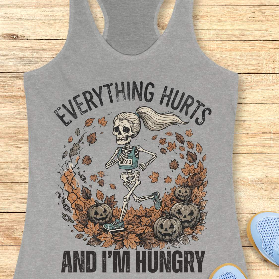 A heather gray racerback tank top laid flat on a light wood background, featuring a Halloween graphic of a skeleton runner surrounded by autumn leaves and pumpkins, with the text 'EVERYTHING HURTS AND I'M HUNGRY'.