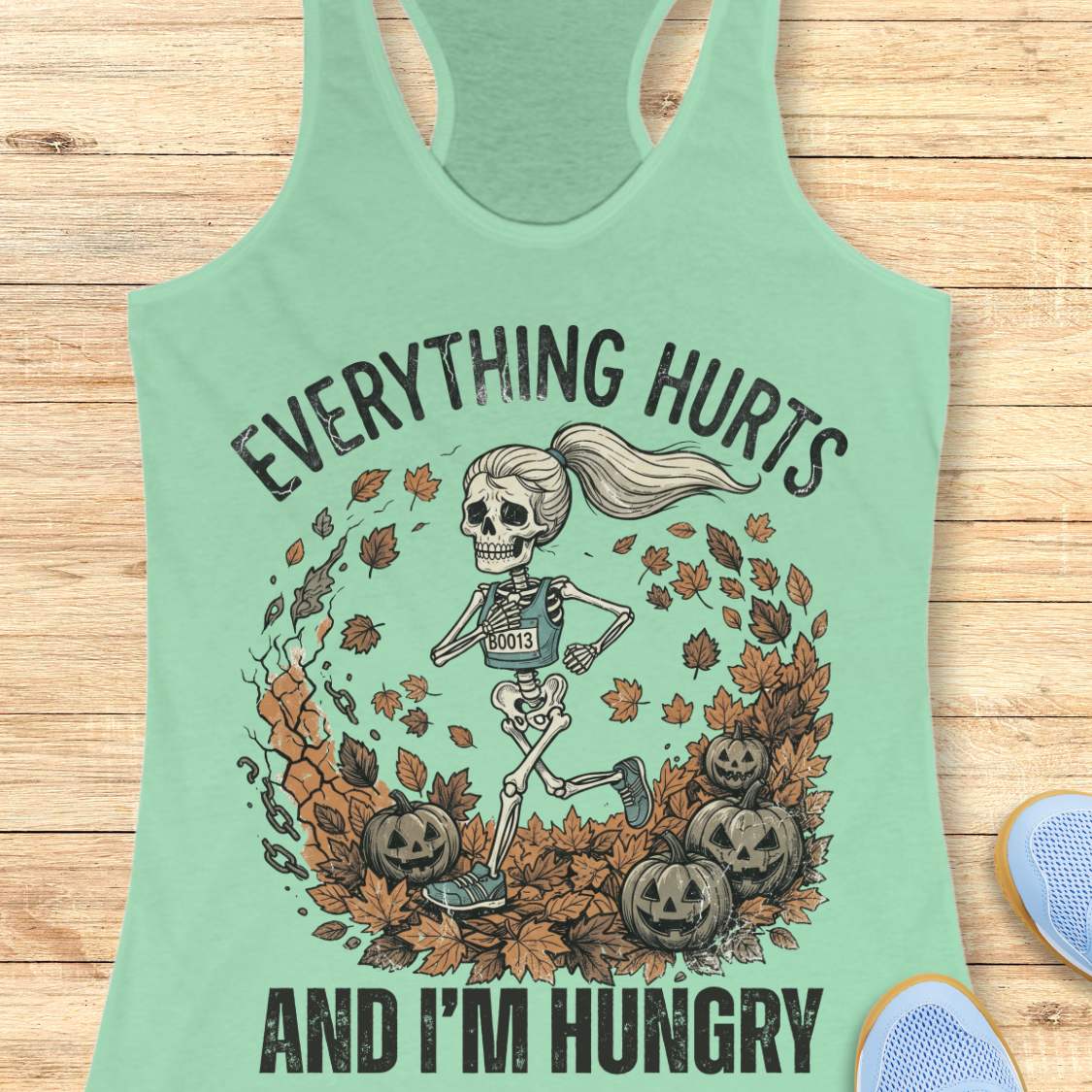 A mint green racerback tank top with a Halloween graphic of a skeleton running through autumn leaves and pumpkins, with the text 'EVERYTHING HURTS AND I'M HUNGRY' printed on the front, displayed flat on a light wood background.