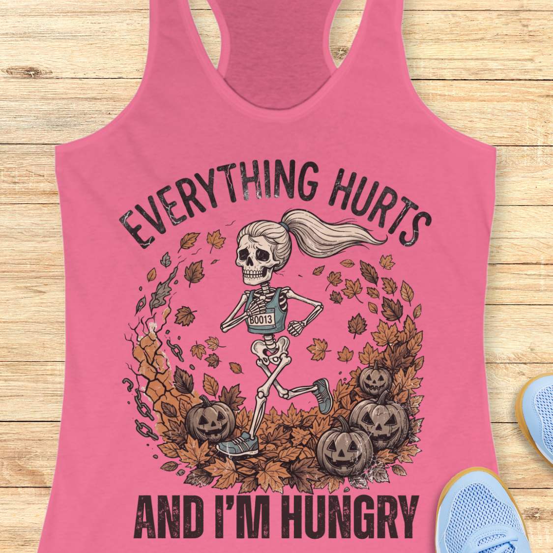 A pink racerback tank top laid flat on a light wood background, featuring a Halloween graphic of a skeleton running through autumn leaves and pumpkins, with the text 'EVERYTHING HURTS AND I'M HUNGRY'. Partial blue shoes are visible at the edges.