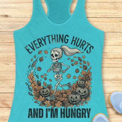 A turquoise racerback tank top laid flat on a light wood surface, featuring a Halloween graphic of a skeleton running through autumn leaves with pumpkins, plus the text 'EVERYTHING HURTS AND I'M HUNGRY'. A partial blue shoe is visible at the lower right edge.