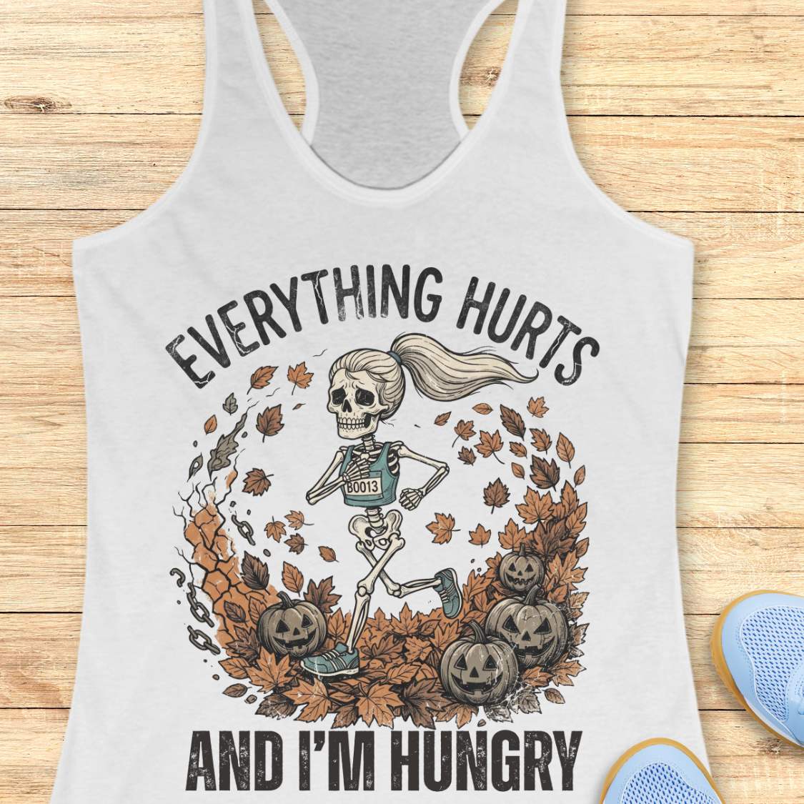 A white racerback tank top laid flat on a light wood background, featuring a Halloween graphic of a skeleton running through autumn leaves with pumpkins, plus the text 'EVERYTHING HURTS AND I'M HUNGRY'. Partial blue shoes are visible at the bottom right.