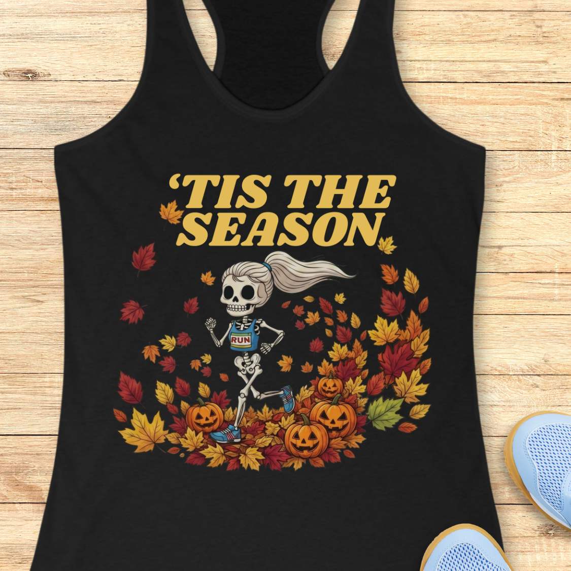 A black racerback tank top laid flat on a light wood surface, featuring a Halloween graphic of a skeleton running through autumn leaves with the text “'TIS THE SEASON” in dark red. A partial light blue shoe is visible at the bottom right corner.