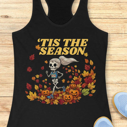 A black racerback tank top laid flat on a light wood surface, featuring a Halloween graphic of a skeleton running through autumn leaves with the text “'TIS THE SEASON” in dark red. A partial light blue shoe is visible at the bottom right corner.
