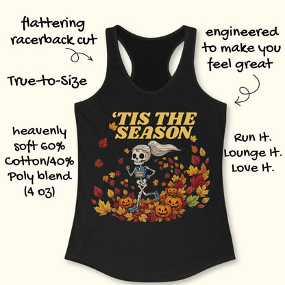 A black racerback tank top laid flat on a light wood surface, featuring a Halloween graphic of a skeleton running through autumn leaves with the text “'TIS THE SEASON” in dark red. A partial light blue shoe is visible at the bottom right corner.