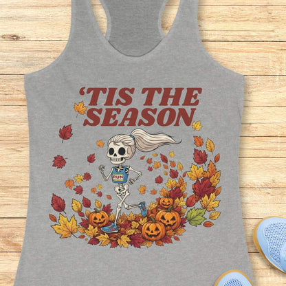 A gray racerback tank top laid flat on a light wood surface, featuring a Halloween graphic of a skeleton running through autumn leaves with the text “'TIS THE SEASON” in dark red. A partial light blue shoe is visible at the bottom right corner.