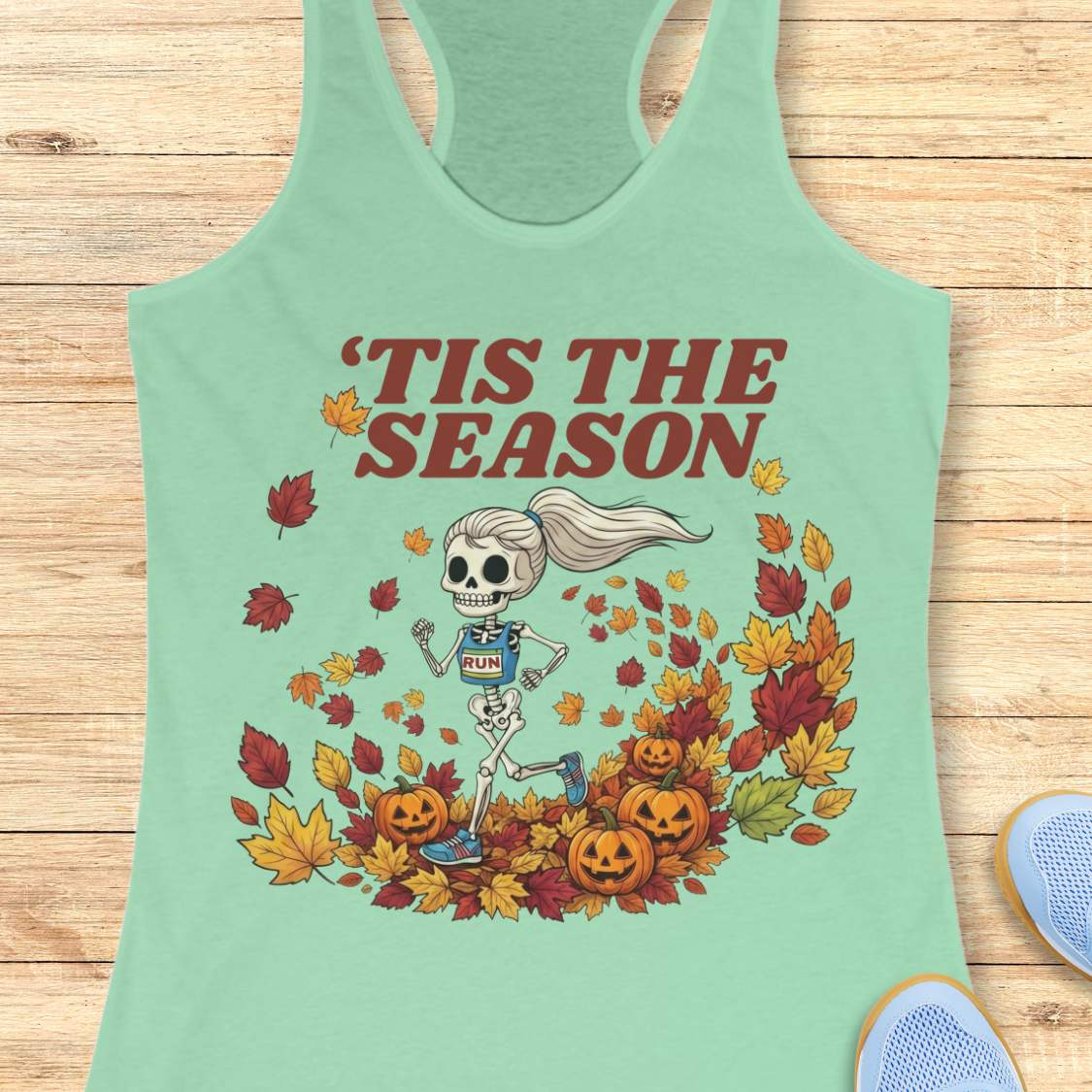 A mint racerback tank top laid flat on a light wood surface, featuring a Halloween graphic of a skeleton running through autumn leaves with the text “'TIS THE SEASON” in dark red. A partial light blue shoe is visible at the bottom right corner.