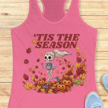 A pink racerback tank top laid flat on a light wood background, featuring a Halloween graphic of a skeleton runner surrounded by autumn leaves and pumpkins, with the text “'TIS THE SEASON” in dark red. A blue shoe is partially visible at the bottom right.