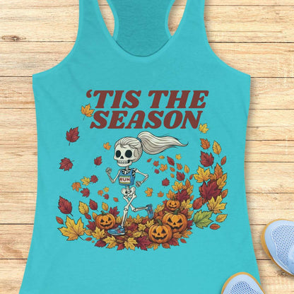 A turquoise racerback tank top laid flat on a light wood surface, featuring a Halloween graphic of a skeleton running through autumn leaves with the text “'TIS THE SEASON” in dark red. A partial light blue shoe is visible at the bottom right corner.