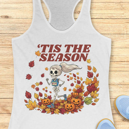 A white  racerback tank top laid flat on a light wood surface, featuring a Halloween graphic of a skeleton running through autumn leaves with the text “'TIS THE SEASON” in dark red. A partial light blue shoe is visible at the bottom right corner.