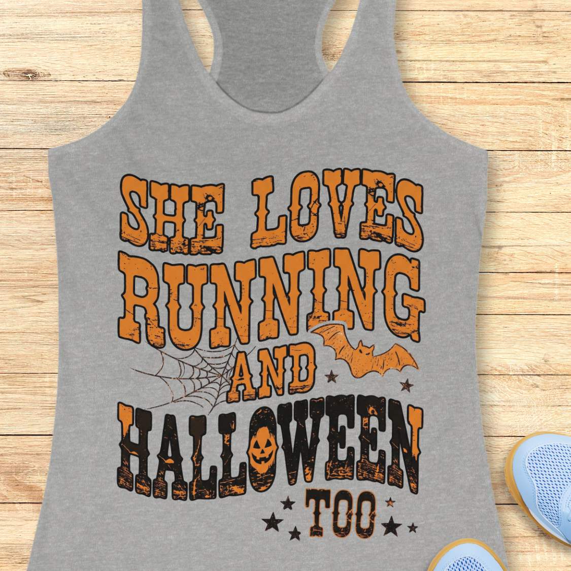 A gray racerback tank top laid flat on a light wood plank background, featuring a large Halloween-themed graphic that reads 'SHE LOVES RUNNING AND HALLOWEEN TOO' in orange and black distressed lettering, with spiderwebs, bats, and stars.