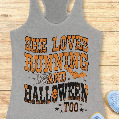 A gray racerback tank top laid flat on a light wood plank background, featuring a large Halloween-themed graphic that reads 'SHE LOVES RUNNING AND HALLOWEEN TOO' in orange and black distressed lettering, with spiderwebs, bats, and stars.