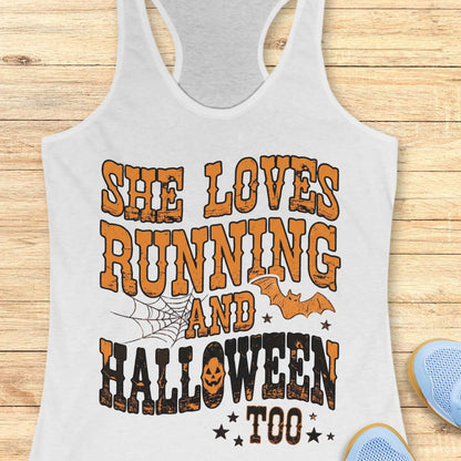 A white racerback tank top laid flat on a light wood plank background, featuring a large Halloween-themed graphic that reads 'SHE LOVES RUNNING AND HALLOWEEN TOO' in orange and black distressed lettering, with spiderwebs, bats, and stars.