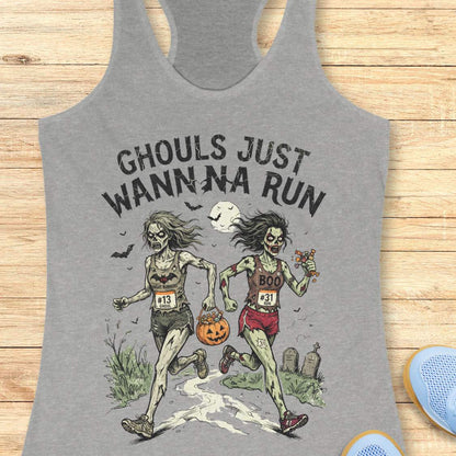 A grey racerback tank top laid flat on a light wood surface, featuring a Halloween graphic of two zombie runners with the text 'GHOULS JUST WANNNA RUN' and a jack-o'-lantern. Part of a blue shoe is visible at the bottom right.
