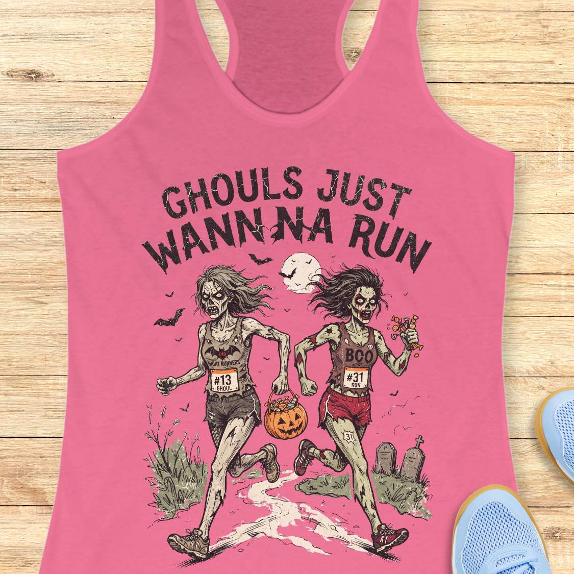 A pink racerback tank top laid flat on a light wood surface, featuring a Halloween graphic of two zombie runners with the text 'GHOULS JUST WANNNA RUN' and a jack-o'-lantern. Part of a blue shoe is visible at the bottom right.