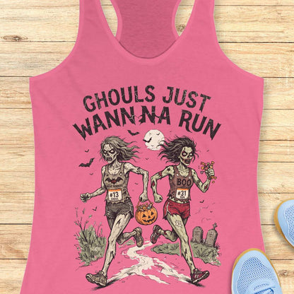 A pink racerback tank top laid flat on a light wood surface, featuring a Halloween graphic of two zombie runners with the text 'GHOULS JUST WANNNA RUN' and a jack-o'-lantern. Part of a blue shoe is visible at the bottom right.