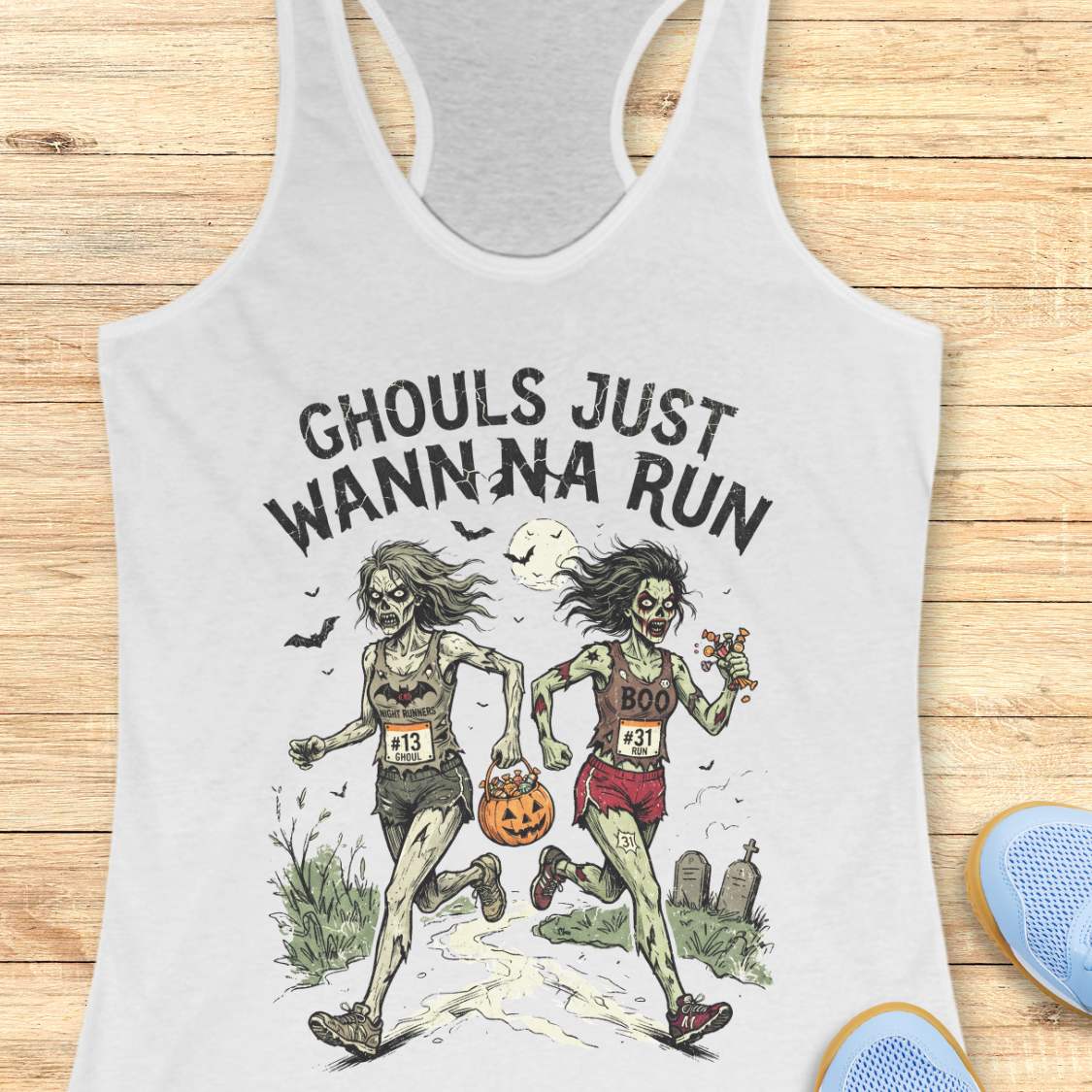 A white racerback tank top laid flat on a light wood surface, featuring a Halloween graphic of two zombie runners with the text 'GHOULS JUST WANNNA RUN' and a jack-o'-lantern. Part of a blue shoe is visible at the bottom right.