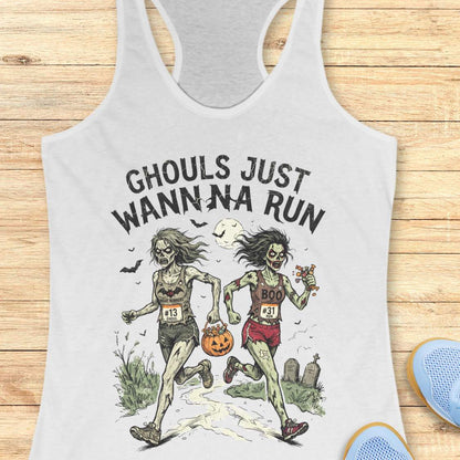 A white racerback tank top laid flat on a light wood surface, featuring a Halloween graphic of two zombie runners with the text 'GHOULS JUST WANNNA RUN' and a jack-o'-lantern. Part of a blue shoe is visible at the bottom right.