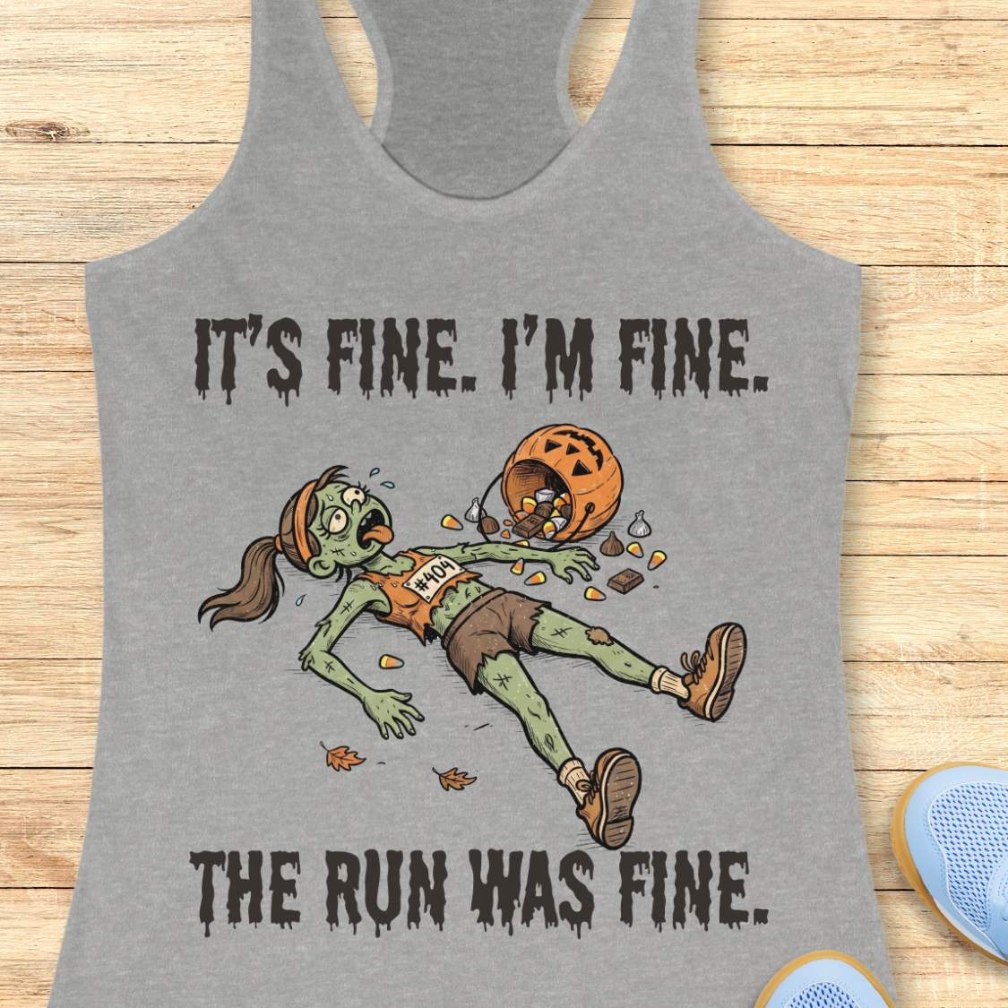 A heather gray racerback tank top laid flat on a light wood surface, featuring a zombie running graphic with the text 'IT'S FINE. I'M FINE.
