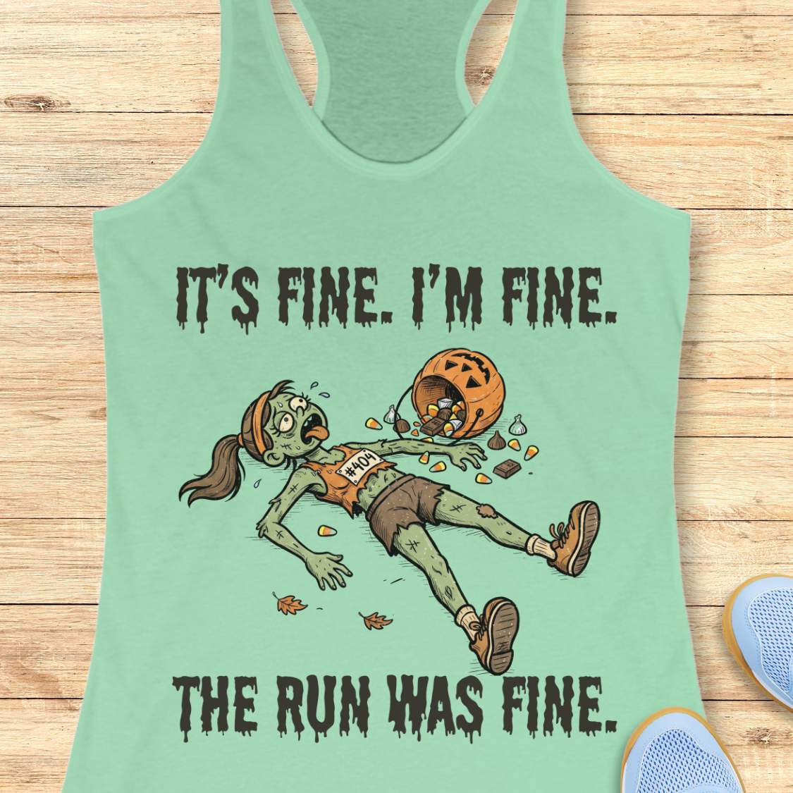 A mint racerback tank top laid flat on a light wood surface, featuring a zombie running graphic with the text 'IT'S FINE. I'M FINE.