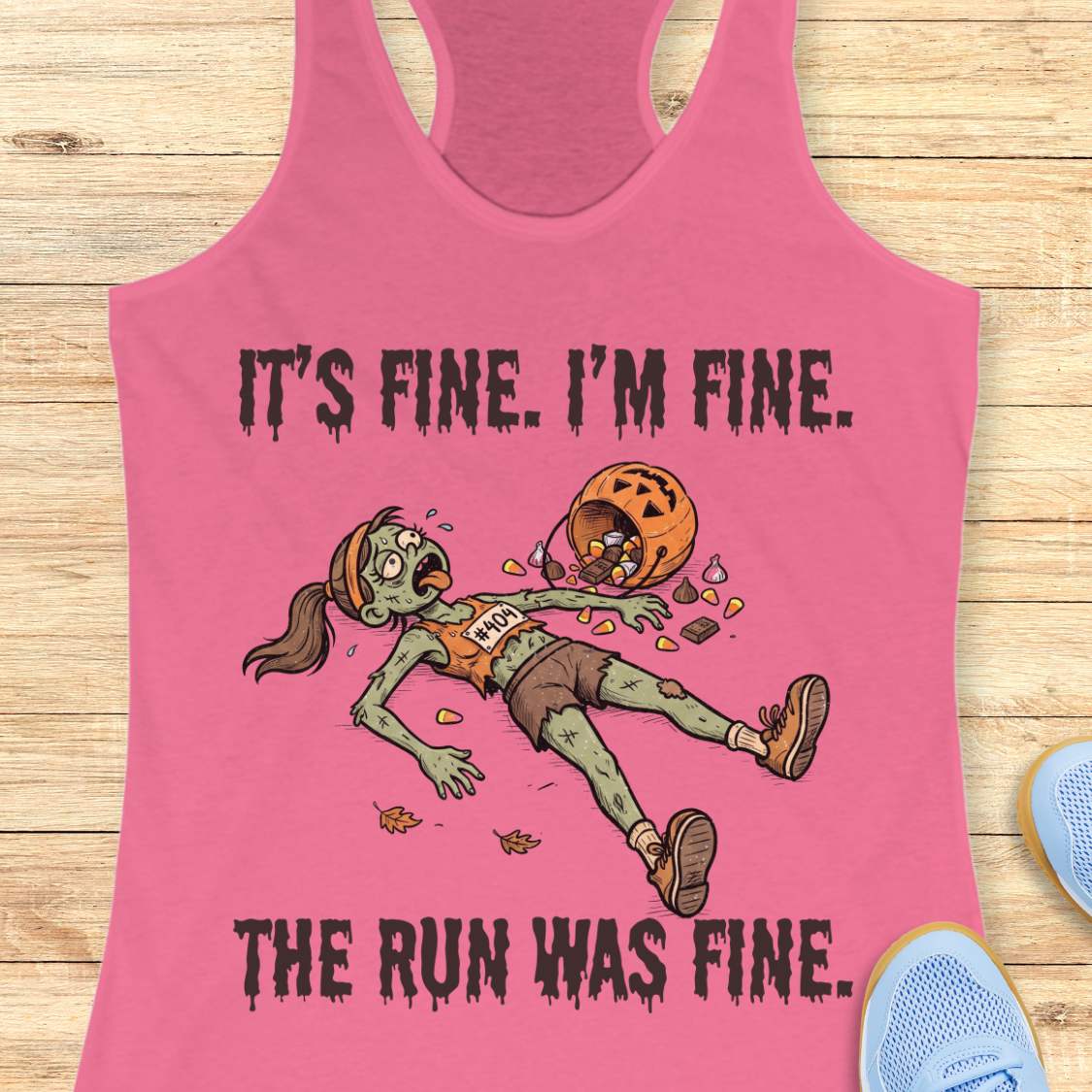 A pink racerback tank top laid flat on a light wood surface, featuring a zombie runner graphic with the text 'IT'S FINE. I'M FINE.