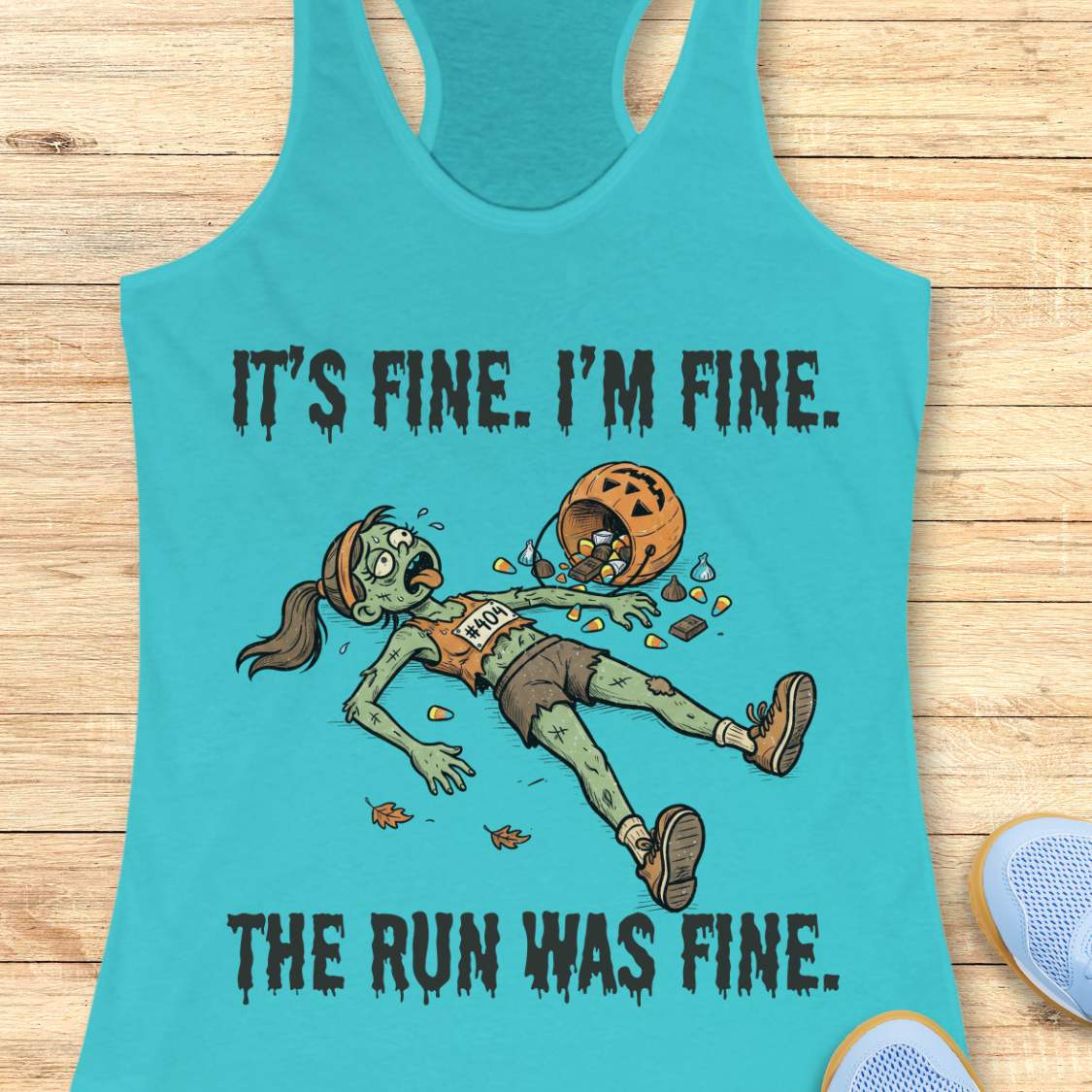 A blue racerback tank top laid flat on a light wood surface, featuring a zombie running graphic with the text 'IT'S FINE. I'M FINE.